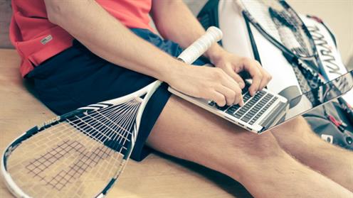 A close-up photograph of a person holding a squash racquet while using a laptop.