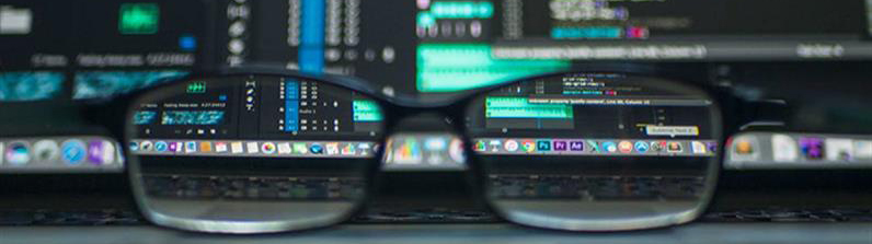 A partial photograph of some reading glasses on a table in the foreground with computer screens in the background.
