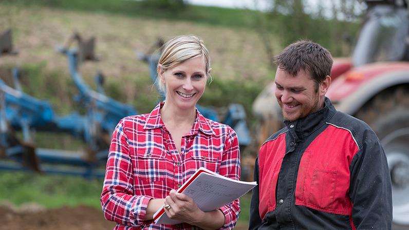 A photograph of a lady with a notepad on her hands and a man looking into it at a farm
