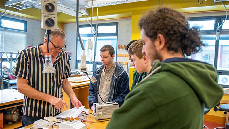 An image of the Electrical Engineering class where students are carefully listing to the lecturer in front of a table with wires, tools and drawing on it