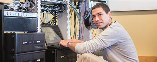 A photograph of a man using a laptop next to networking equipment