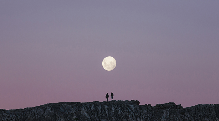 moon in centre of purple blue sky with two people on cliff